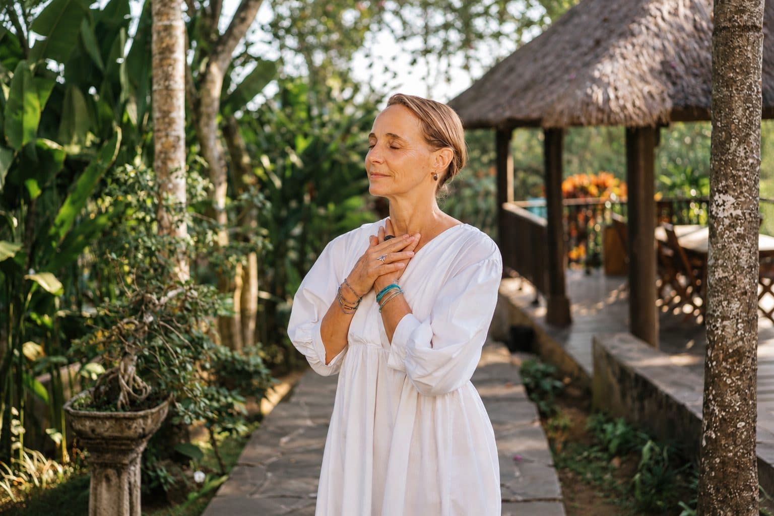 Christiane steht im Garten vor einem Pavillon mit Reetdach, die Hände am Herzen.